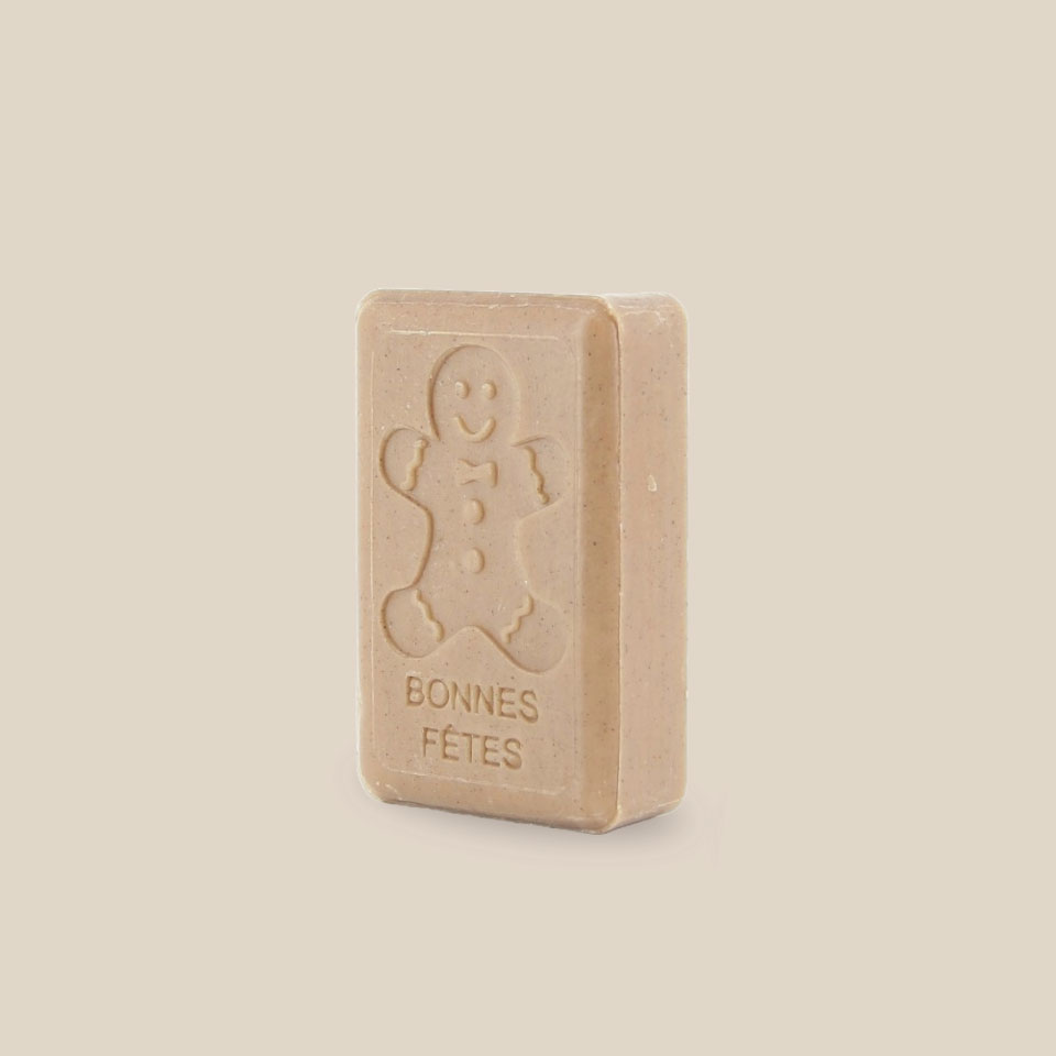 Photo produit prise de côté : Savonnette marseillaise au beurre de karité, de couleur brune pâle avec petit grain, parfumée et estampillée Bonnes fêtes accompagnée d'un bonhomme pain d'épices. Fabriqué à Marseille. 125g.