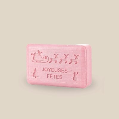 Photo produit prise de côté : Savonnette marseillaise au beurre de karité, de couleur rose, parfumée et estampillée Joyeuses fêtes accompagnée du traineau du père Noel, sapin et bonhomme de neige. Fabriqué à Marseille. 125g.